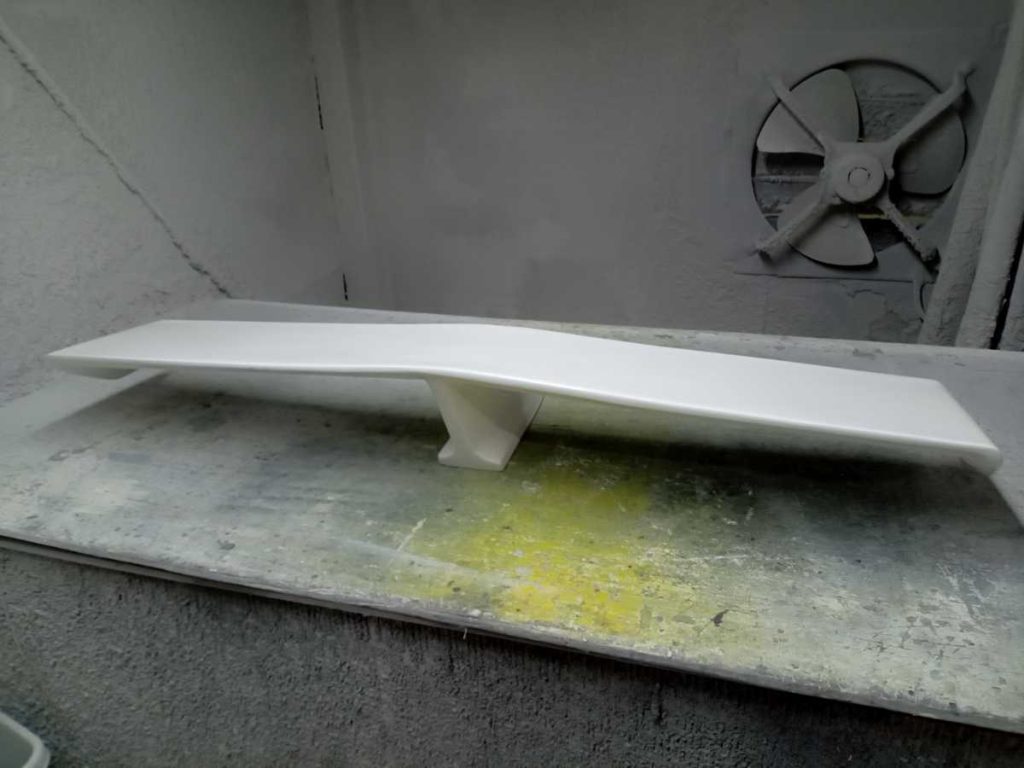 White car spoiler in a spray booth during painting process, with ventilation fan visible in the background.