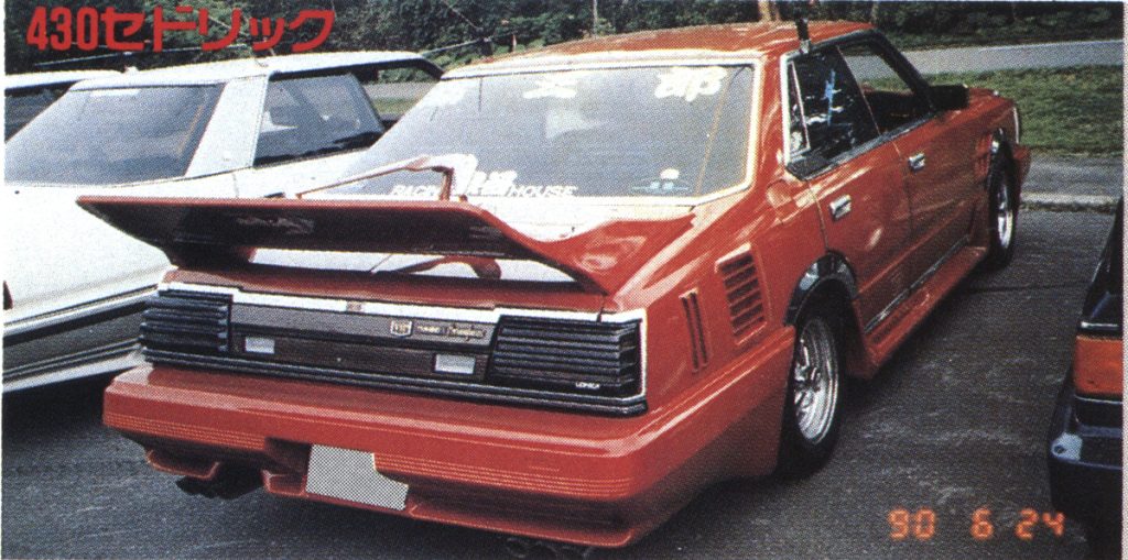 Rear view of a modified red 430 Cedric car, with a large rear spoiler, parked next to other vehicles.