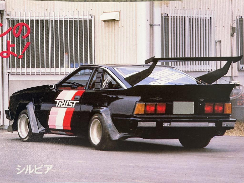 Custom black Nissan Silvia with large rear wing and TRUST decal in front of industrial building in Japan.