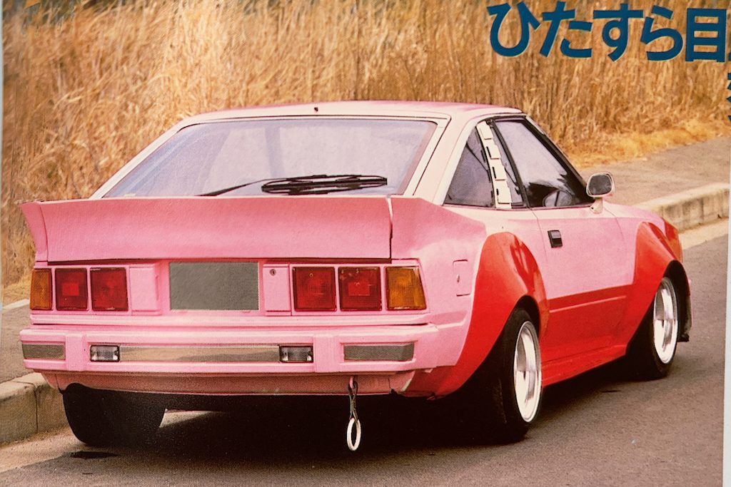 Rear view of a pink modified Nissan Gazelle S110 car parked by the roadside, featuring wide body kit and spoiler. Japanese text in upper right.