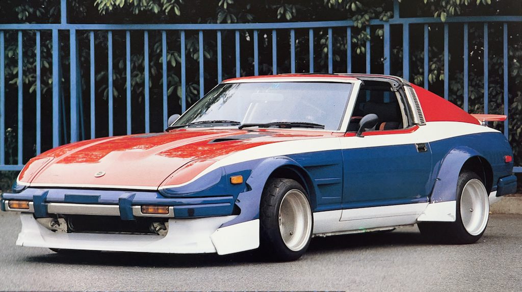 A retro Nissan Fairlady Z S130 with a red, white, and blue color scheme and Nissan Fairlady Z S30 Works fender parked in front of a blue fence with greenery in the background.