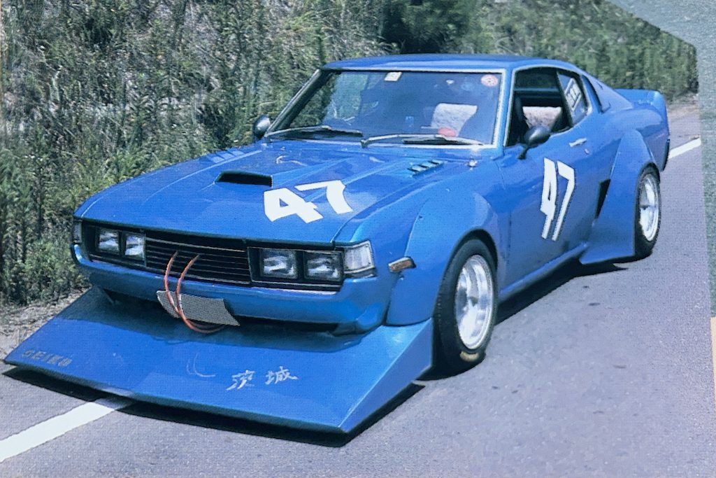 Modified blue race car with number 47, front bumper text: Seiko, Japan. Driving on a road with greenery in the background.