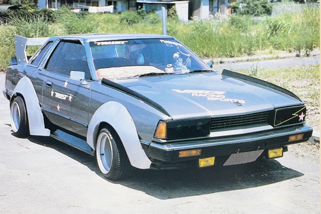 Customized Nissan Silvia S110 with wide LB works fenders, spoiler, and Trust and Racing Sports decals, parked outdoors.