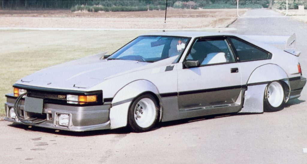 Customized retro Japanese Toyota Celica XX car with wide Savanna works fenders and a low stance parked on a rural road with green fields in the background.