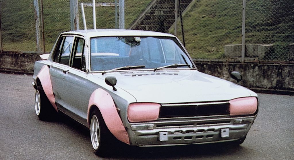 Vintage modified car with pink headlight covers and fender flares parked near a fence on an overcast day.