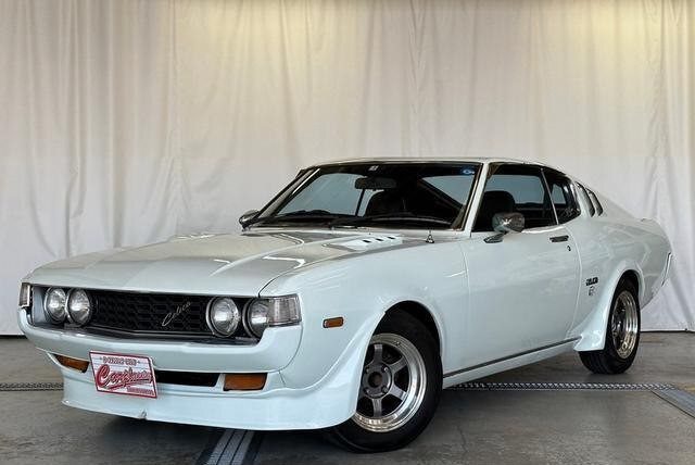 Classic white Toyota Celica GT in showroom, front side view, with Japanese license plate.