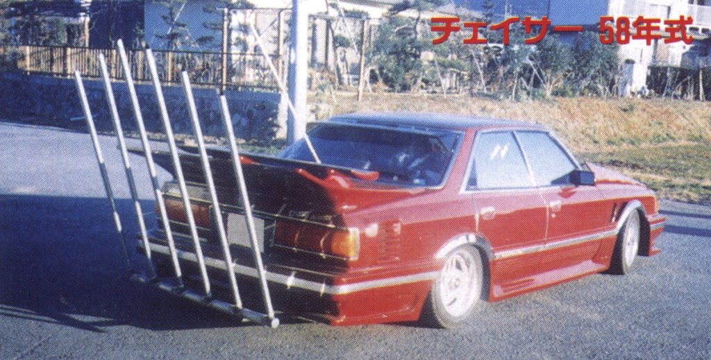 Modified red car with extended exhaust pipes and kanji text reading Chaser 58-year model on a sunny street in Japan.