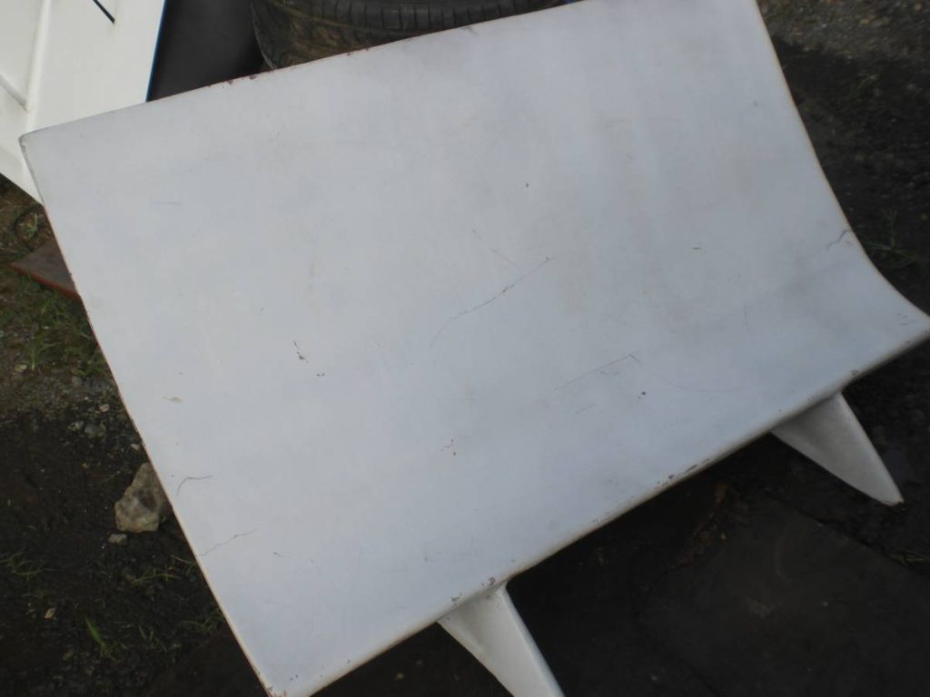 An old, white car part with visible cracks and scratches, lying on the ground next to a tire and other items.