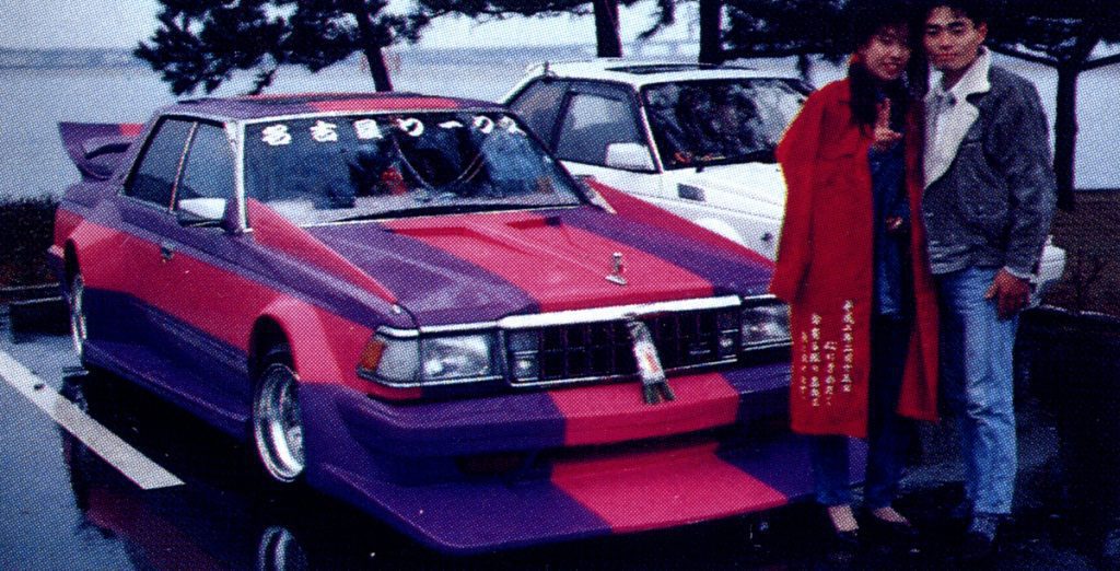 A couple standing next to a vibrantly colored, modified car with Japanese text on its windshield and clothing, in a parking lot.