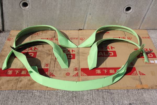 Green Nissan Fairlady Z S30 Works fender car body kit parts on cardboard laid on a concrete floor.