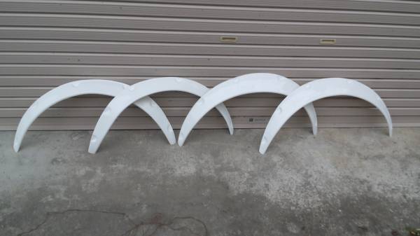 Four white car fender flares arranged against a garage door background, ideal for vehicle modifications and styling.