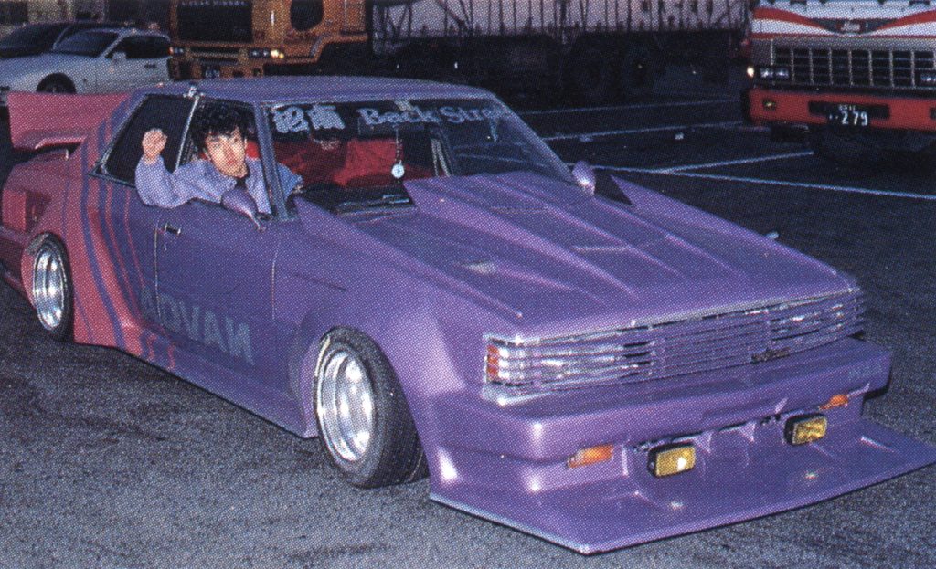 A man in a custom purple car with Back Step and 改造車 (custom car) decals in a parking lot at night.