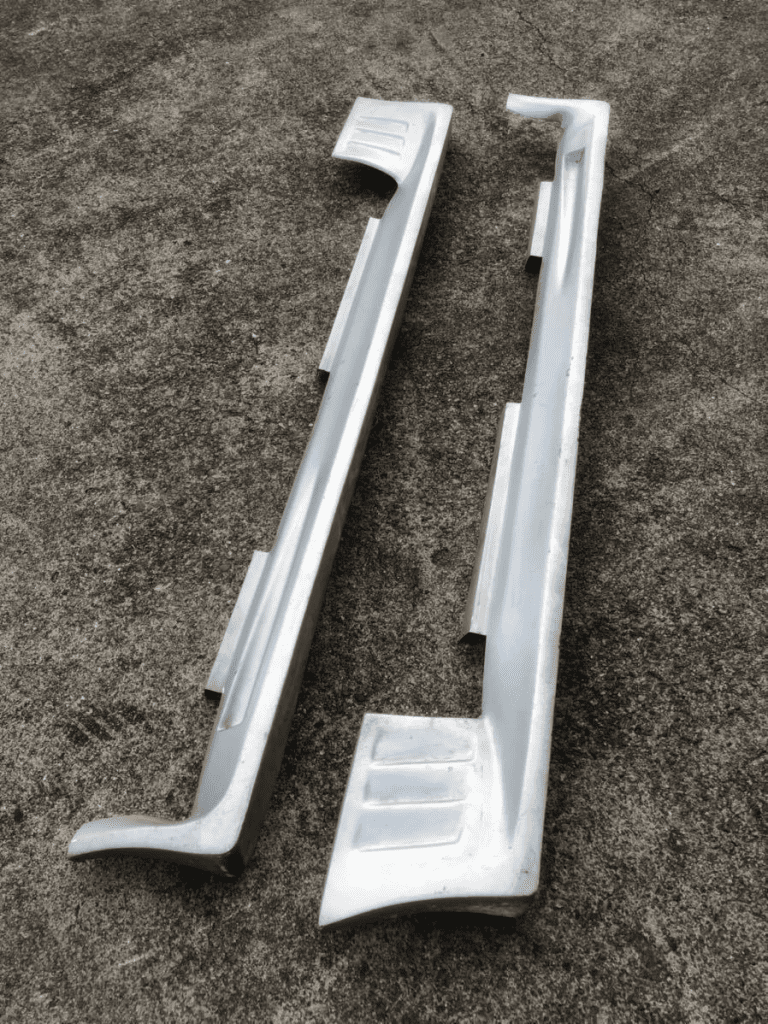 Two white car side skirts on grey concrete, ready for vehicle installation or replacement, automotive parts