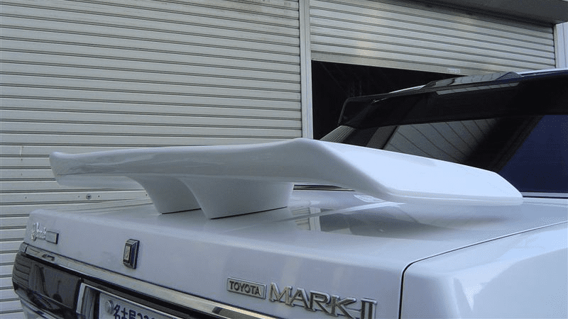 Custom rear spoiler on a white Toyota Mark II car parked by a closed shutter door.