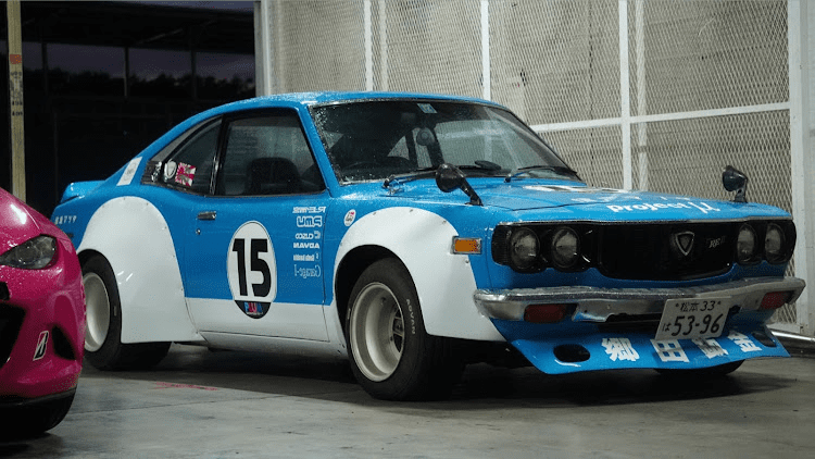 Vintage blue and white Mazda RX-3 Savanna with works over fender with number 15 on the side, Japanese license plate, parked in a garage.