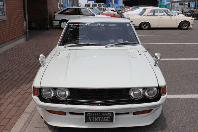 Front view of a classic white vintage car parked in a lot with Japanese text on the windshield and Classic House Vintage plate.