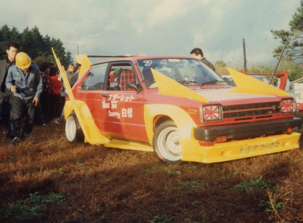 Modified Toyota Starlet with custom yellow and red body at a Japanese car event. Japanese text reads Starlet and Toyota Works.