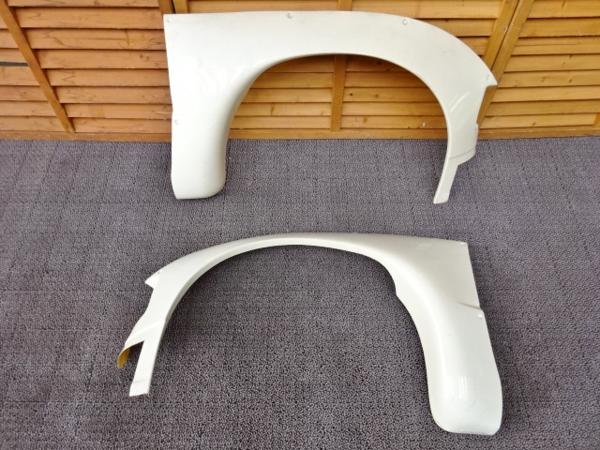 Two white car fenders placed on a gray carpet, with a wooden panel background. Ideal for vehicle spare parts.