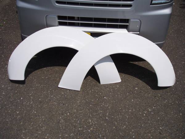 White fender flares in front of a grey vehicle fascia, showcasing automotive parts on an asphalt surface.