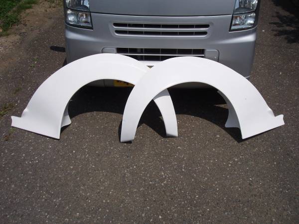 Two large white fender flares placed in front of a silver van on a paved ground outdoors.