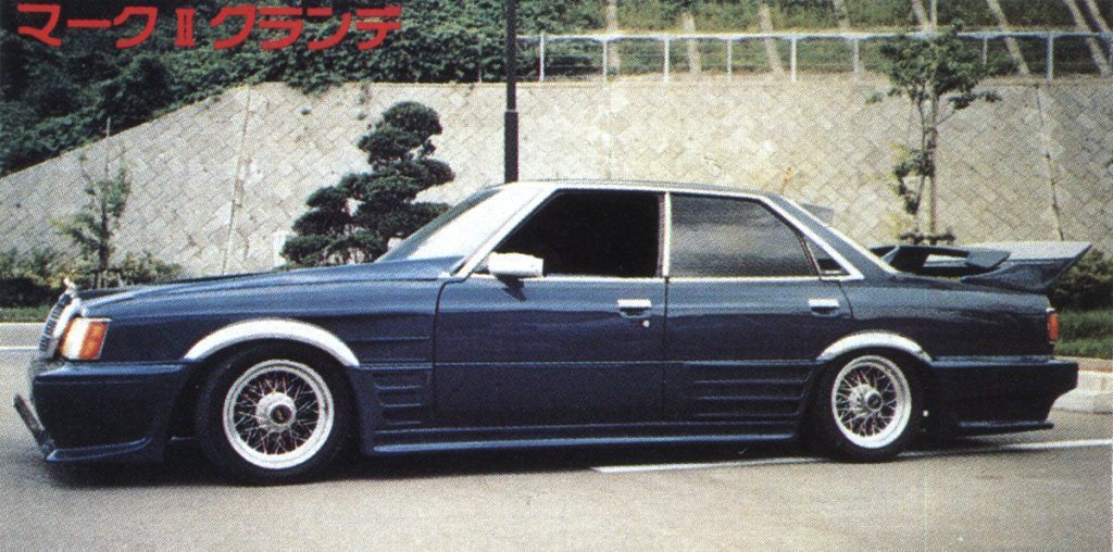 Modified retro Toyota Mark II Grande with aerodynamic body kit and aftermarket rims, parked against a garden backdrop.