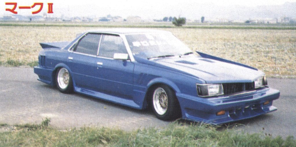 Blue modified Toyota Mark II parked in countryside, showing off wide body kit and custom wheels.