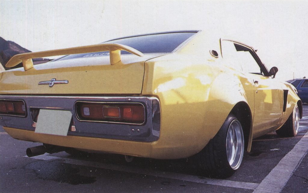 Rear view of a vintage yellow Toyota Celica parked, showcasing its distinctive design and rear spoiler.