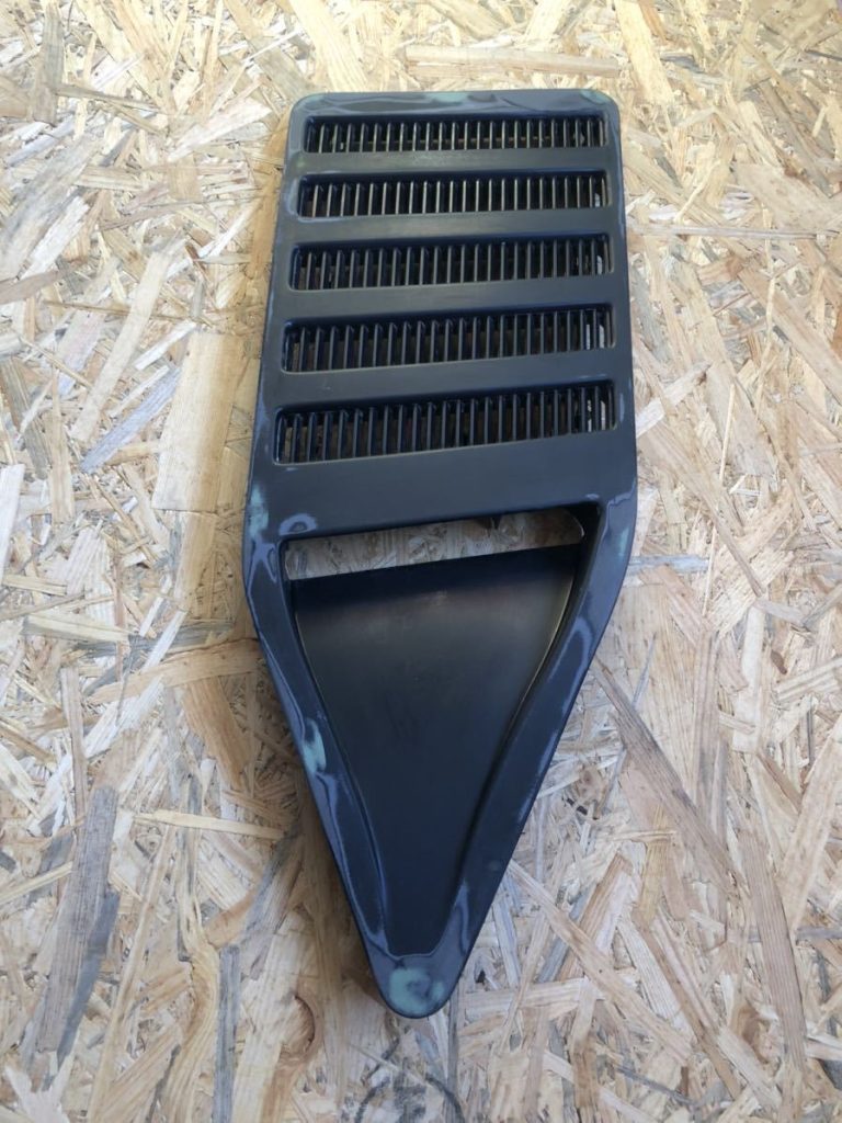 Black automotive vent grille for hood or bumper on plywood background.