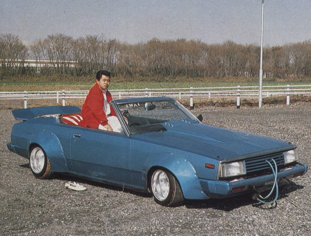 A man in a red jacket sits in a modified blue convertible car with a large spoiler in an outdoor setting.