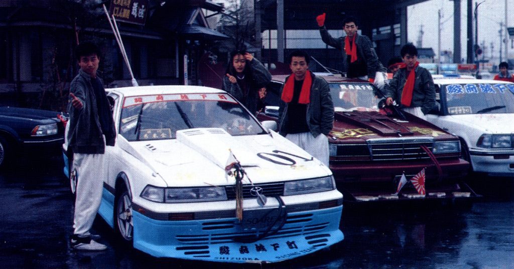 Group of five men posing with custom cars in a Japanese street with banners and signs written in Japanese characters.