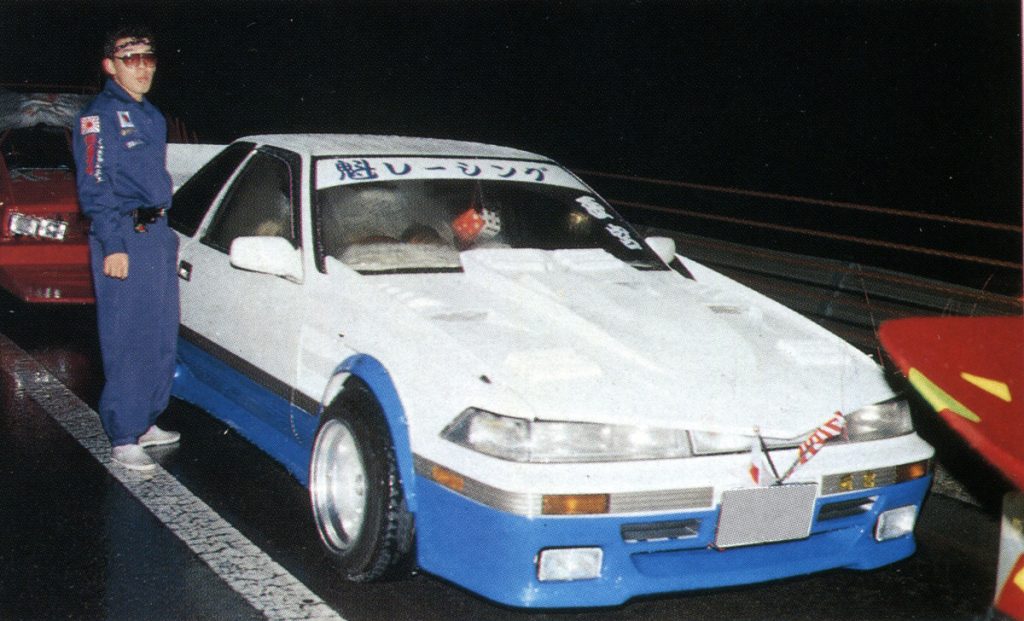 Street racer stands beside white and blue modified car at night; Japanese text on car reads 魅レーシング meaning 'Charm Racing'.