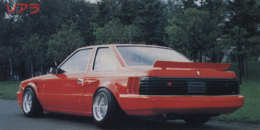 A classic red Toyota Soarer sports car with a spoiler, parked outdoors surrounded by lush greenery.