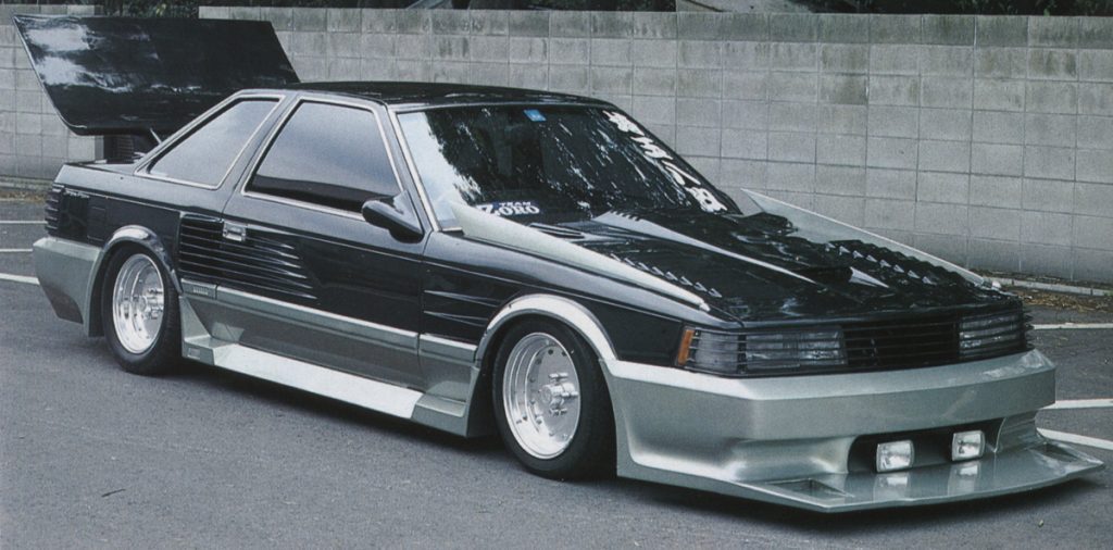 Modified vintage car with aggressive body kit and custom detailing, parked on a street with concrete wall background.