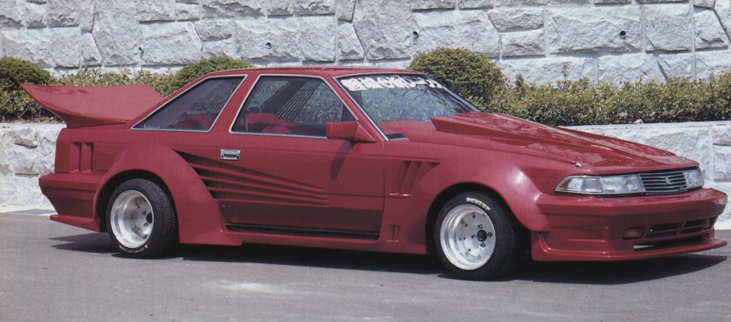 Wide-body red sports car with large rear spoiler and decals, parked outdoors in front of a stone wall.