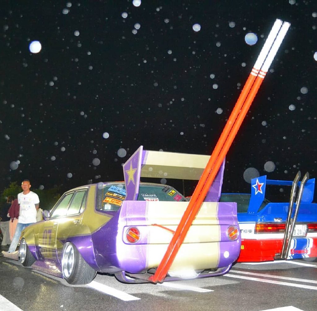 Modified cars on display at a night car meet in Japan, including a purple car with large chopsticks decoration.