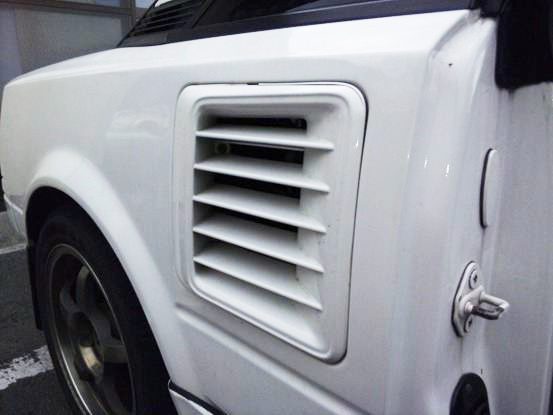 Close-up of a white car’s side air vent, showcasing classic design with slotted vent cover and rear wheel.