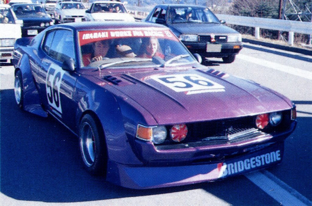 Vintage purple racing car with number 58 and Bridgestone branding on a highway, followed by other vehicles.