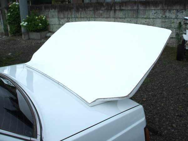 Rear view of a white vehicle's trunk lid slightly open in an outdoor setting with greenery and a concrete wall in the background.
