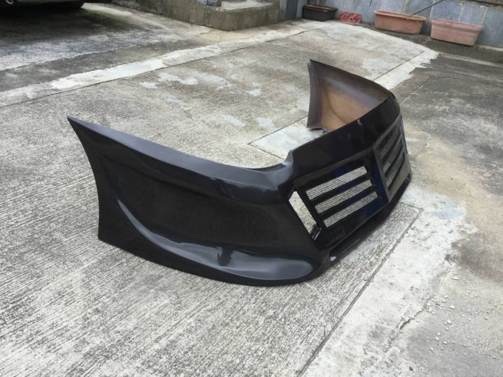 Black car front bumper placed on concrete ground, clear view of grill design, surrounding planters in the background.