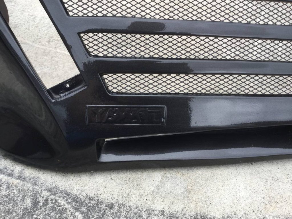 Close-up of a black car grille with Mazda logo and mesh pattern on a concrete surface.