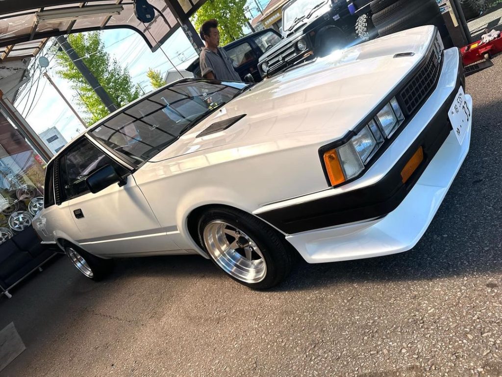 White classic car with black trim and polished wheels in a showroom, person in the background, Japanese license plate.