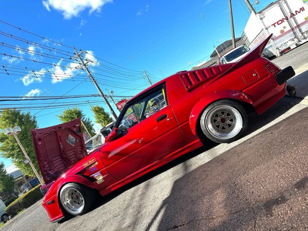 Custom red modified car with hood open, under a clear blue sky. Truck in the background reads TONAMI in Japanese letters.