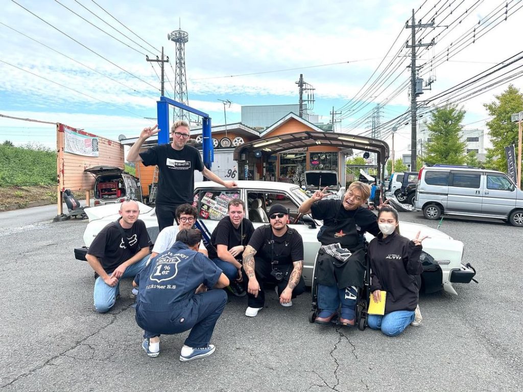 A group of people pose in front of a garage and car, some making peace signs, featuring stickers and a 1%er jacket.