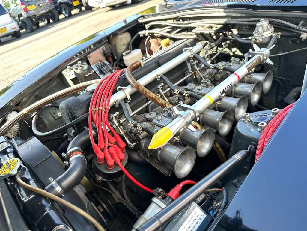Close-up of a high-performance car engine with visible red ignition wires and 秀MAX text on the fuel rail in a parking lot.