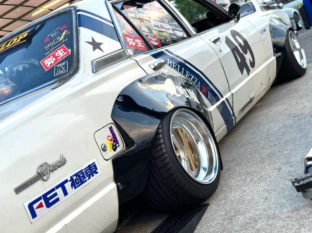 Modified Japanese car with race stickers, showcasing custom rims and a lowered suspension. Text reads Grande and FET.