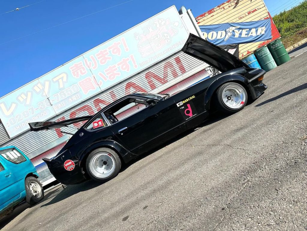 A black modified sports car with an open hood parked outside a garage with Japanese signage in the background.