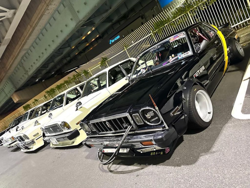 A lineup of retro Japanese cars under bridge; captions in Japanese read, 選手一家, meaning Racer Family.