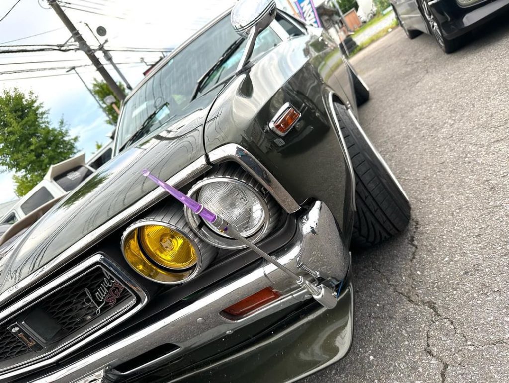 Close-up of a vintage 'Laurel SGX' Japanese car with yellow headlights and a customized hood ornament.