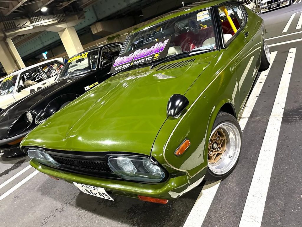 Green classic car with black headlights and golden rims at a car meet. I don’t know the displayed Japanese text.