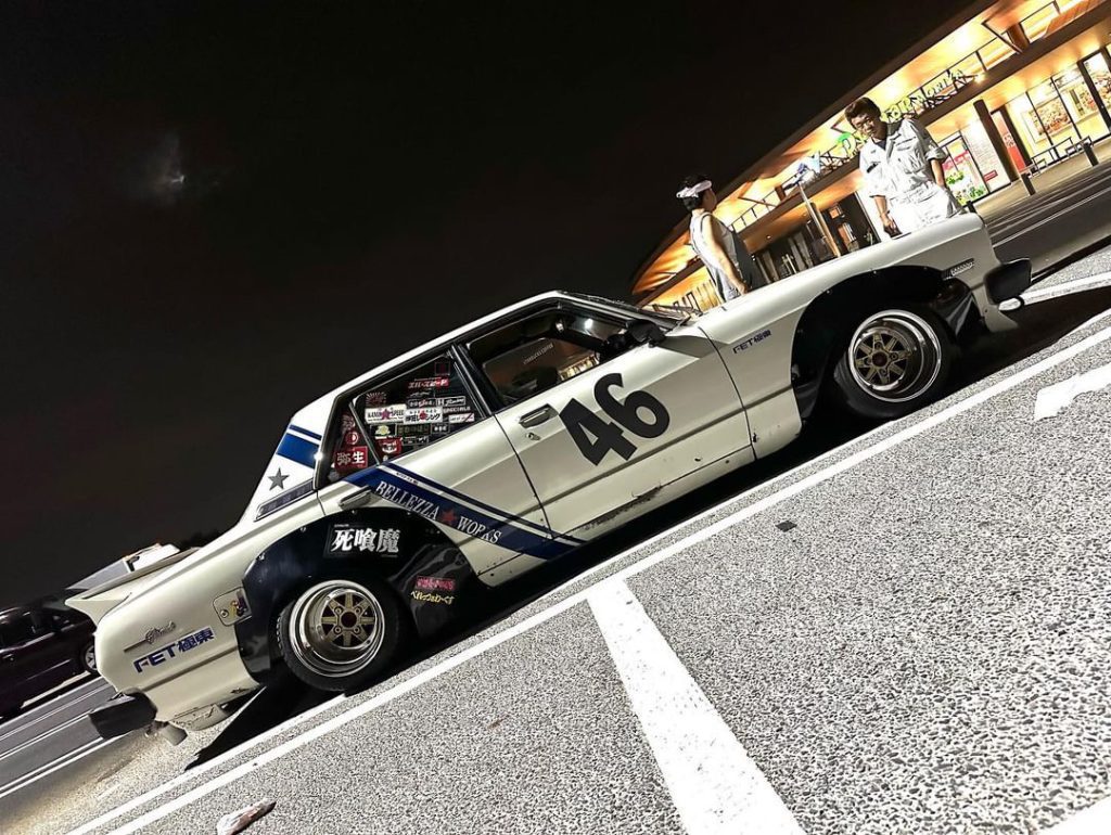 Classic white sports car with a number 46, parked at night, two people standing beside; Japanese shops in the background.